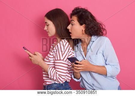 Horizontal Shot Of Two Women Sharing Social Media Videos In Smart Phone Indoors, Attractive Dark Hai