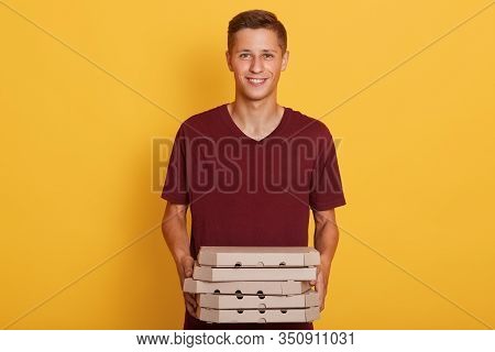 Hirizontal Shot Of Handsome Man Wearing Maroon Casual T Shirt, Holding Pizza Boxes In Hands, Looking
