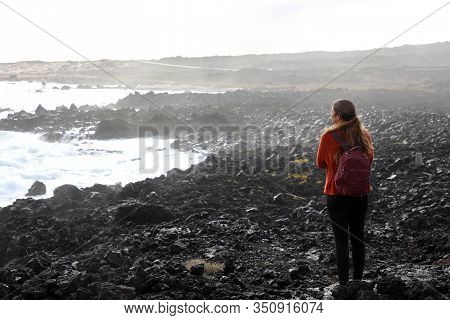 Desolate Volcanic Black Rocks With Lonely Girl. Back View Of Young Woman Standing Reflexive And Sad 