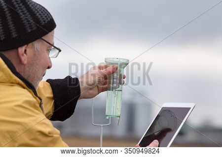 Senior Farmer With Tablet Squatting Beside Rain Gauge In Field