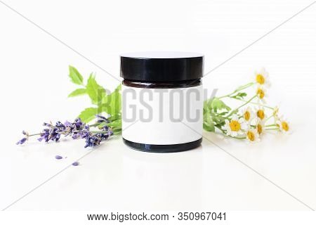 Closeup Of Dark Glass Jar With Blank Paper Label. Mint Leaves, Lavender And Feverfew Flowers On Whit