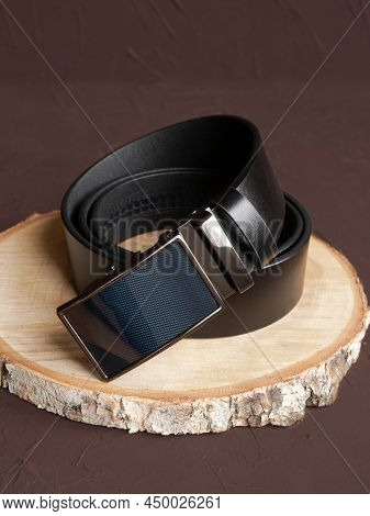Rolled-up Black Fashionable Men's Leather Belt With Automatic Buckle On Wood Slice On Brown Backgrou