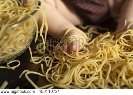 Little Chubby Kids Foot In Pasta On A Wooden Brown Background, Table. Feeding Concept. Spaghetti Sma