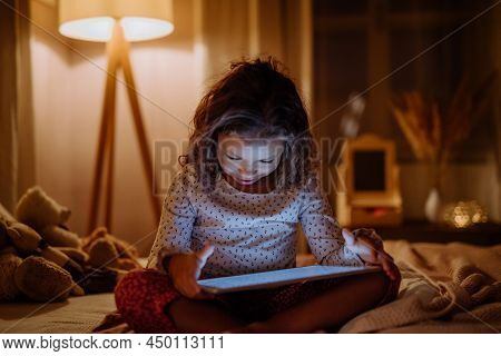 Happy Little Multiracial Girl Using Tablet On Bed In Evening At Home.