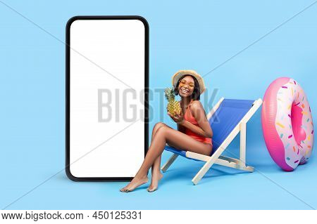 Joyful Young Woman In Bikini Sitting In Lounge Chair Near Giant Smartphone With Blank Screen, Mockup