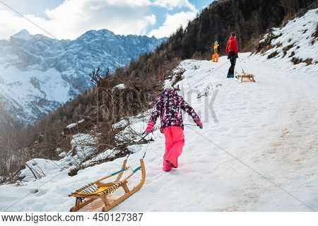 Girl In Colourful Snowsuit Walking Behind Her Dad And Brother, Going Uphill And Pulling A Wooden Sle