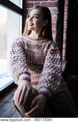 Attractive girl in stylish casial clothes sits on a windowsill looking joyfully out the window in a loft room. Happy and successful people. 