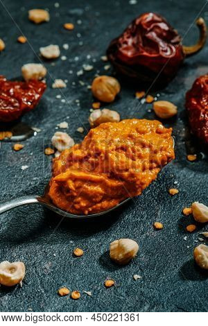 a spoon with some romesco sauce, typical of Catalonia, Spain, next to some of the ingredients to prepare it, such as tomatoes, garlic, nyora pepper or hazelnuts, on a dark surface
