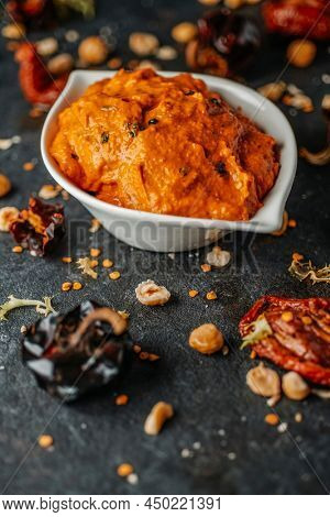 a white ceramic bowl with some romesco sauce, typical of Catalonia, Spain, made with olive oil, garlic, tomato, nyora pepper or hazelnuts, on a dark surface with some of the ingredients to prepare it