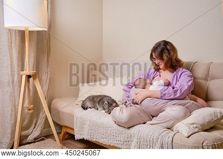 Woman Mother Breastfeeds An Infant Baby Sitting On A Home Sofa. Mom Feeds Milk Toddler Baby In Livin