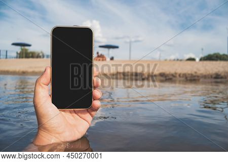 Phone In A Man's Hand. Against The Backdrop Of Water, Beach And Blue Sky.