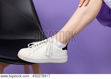 Cropped Photo Of A Young Woman Leg In White Sneekers Putting On A Chair On Purple Background, Health