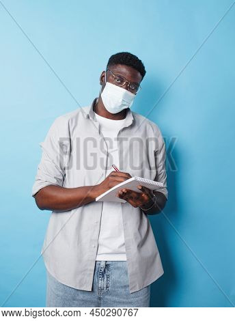 An African-american Man In A Medical Mask Writes Information In A Notebook On An Isolated Blue Backg