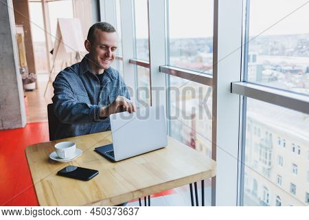Successful Happy Business Man Is Sitting At A Table In A Cafe, Holding A Cup Of Coffee And Using A L