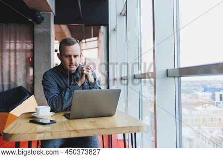 Successful Happy Business Man Is Sitting At A Table In A Cafe, Holding A Cup Of Coffee And Using A L