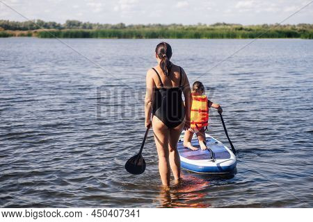 Middle Aged Caucasian Mother Looking After Little Daughter Sup Boarding On Lake With Oar In Hands In