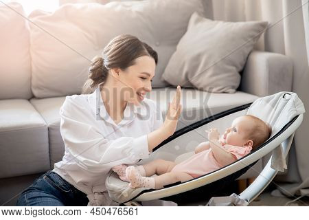 Young Mother Playing With Baby Lies In Automatic Carrycot, Sunlight At Home