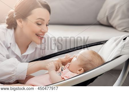Happy Mother Playing With Girl Baby Lies In Automatic Carrycot, Sunlight At Home