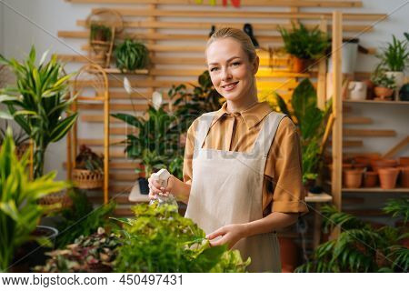Portrait Of Smiling Female Florist In Apron Spraying Water On Houseplants In Flower Pots By Sprayer,