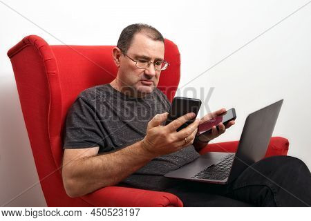 Middle-aged Man With A Laptop On His Lap Sed In A Red Armchair. The Man Is Holding Two Smartphones