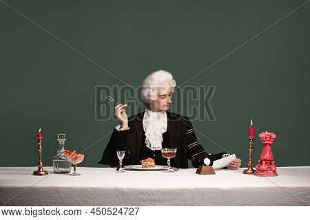 Portrait Of Young Elegant Man In Peruke And Vintage Jacket Sitting At Table Isolated On Dark Green B