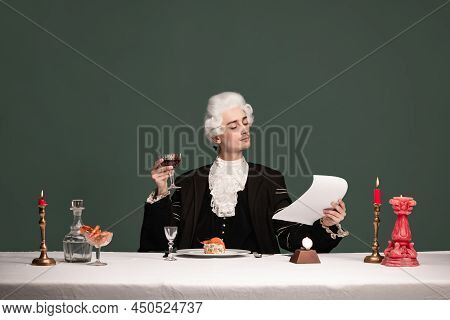 Portrait Of Young Elegant Man In Peruke And Vintage Jacket Sitting At Table Isolated On Dark Green B