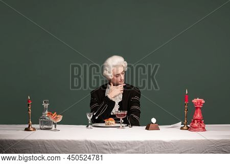 Portrait Of Young Elegant Man In Peruke And Vintage Jacket Sitting At Table Isolated On Dark Green B