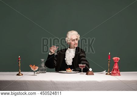Portrait Of Young Elegant Man In Peruke And Vintage Jacket Sitting At Table Isolated On Dark Green B