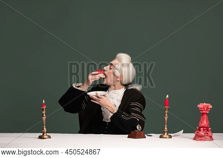 Portrait Of Young Elegant Man In Peruke And Vintage Jacket Sitting At Table Isolated On Dark Green B