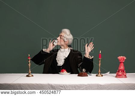 Portrait Of Young Elegant Man In Peruke And Vintage Jacket Sitting At Table Isolated On Dark Green B