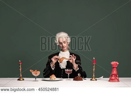 Portrait Of Young Elegant Man In Peruke And Vintage Jacket Sitting At Table Isolated On Dark Green B