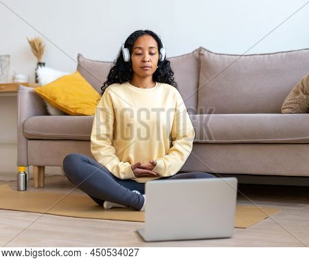 African-american Female Listening To Music In Headphones While Meditating With Laptop At Home