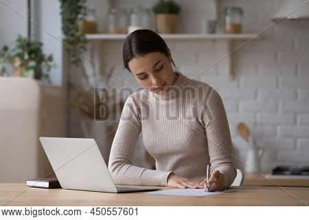 Woman Sit At Table With Laptop Jotting Information On Paper