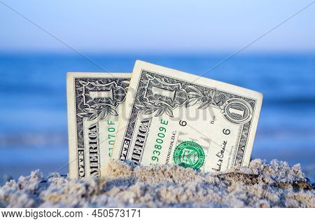 Two One Dollar Bills Half Buried In Sand On Sandy Seashore Close-up.
