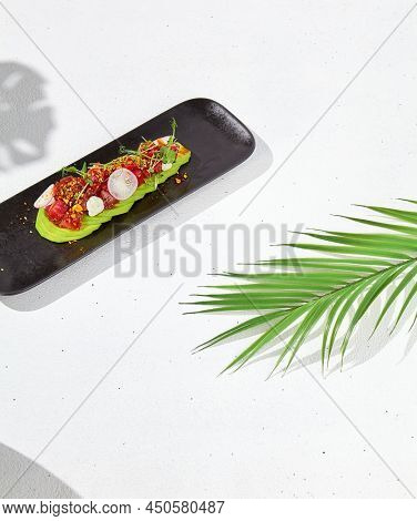 Modern food - tuna tartare on sliced avocado in black plate. Avocado starter with tuna and radish in asian style. Contemporary asian dish. Healthy food with avo and marinated fish on white background