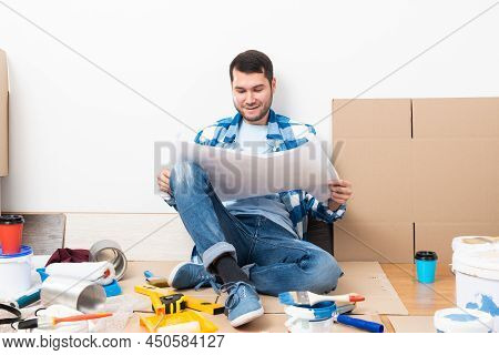 Guy Looking At Paper Blueprint While Sitting Near Wall At Home. Young Man Planning Home Design Proje