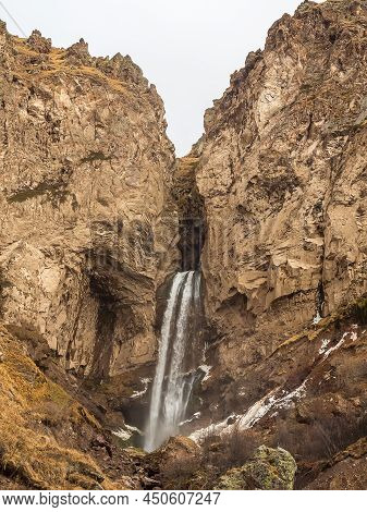 In High Massive Rocky Mountains A Waterfall Flows On A Cloudy Autumn Day.