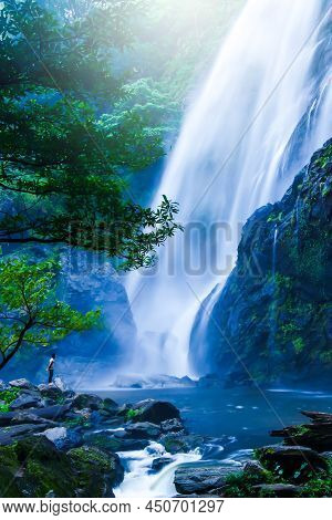 An Asian Barechested Man Stands On The Rock And Looks At The Great Waterfall In Rain Season. Khlong 