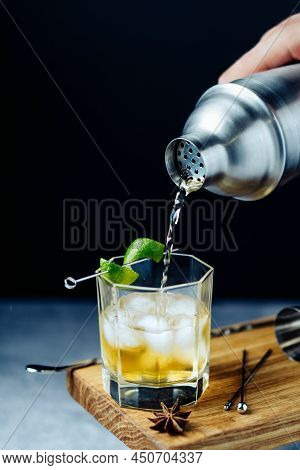 Alcohol Cocktail Pouring From Shaker Into Gless With Lime Peel Ice On Wooden Desk. Party Time