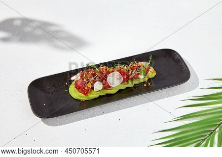 Modern food - tuna tartare on sliced avocado in black plate. Avocado starter with tuna and radish in asian style. Contemporary asian dish. Healthy food with avo and marinated fish on white background