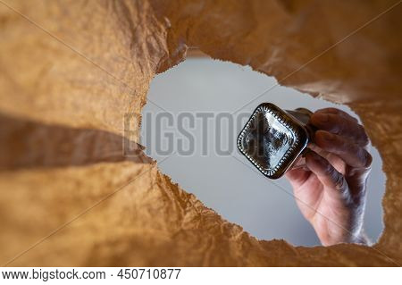 A Hand Tosses A Black Bottle Into A Paper Bag. A Mature Man's Hand Holds The Glass Bottle Over The O