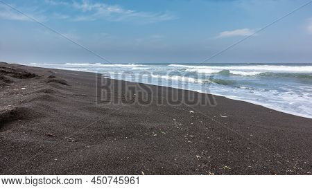 The Coast Of The Pacific Ocean Is Covered With Black Volcanic Sand. Long Turquoise Surf Waves Roll A