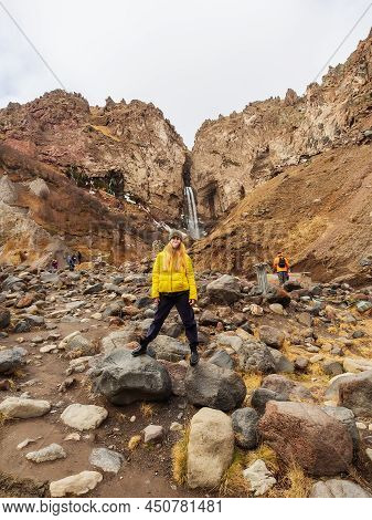 A Woman In A Face Mask Stands On Large Stones In A Rocky Gorge Against The Backdrop Of A Waterfall A