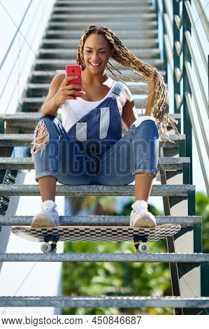Young Black Woman With Coloured Braids, Sitting On Some Steps While Consulting Her Smartphone With H