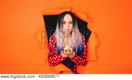 Woman With Toy House Looking Through Hole In Paper. Female With Afro Braids And Miniature Wooden Hou