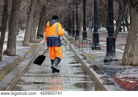 Cleaning Street In A City, Janitor With Shovel Removes Melting Snow In Spring Park. Work Of Housing 