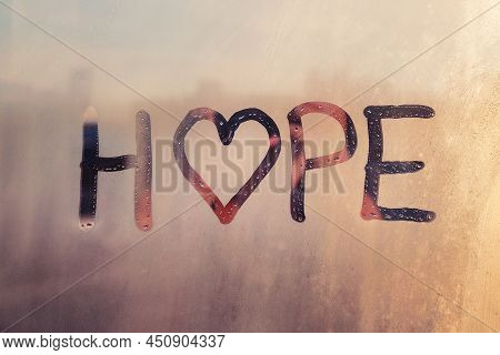 Handwritten Word Hope With Heart Shape On Misted Sunset Glass On Window Flooded With Raindrops