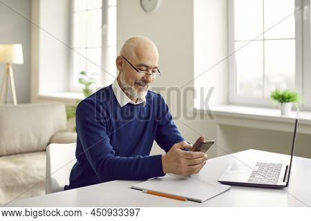 Happy Senior Man Using His Mobile Phone While Sitting At Table With Laptop Computer