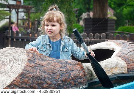 A Little Girl With Wavy Blond Hair In An Amusement Park Is Sailing On A Boat, She Has An Oar In Her 