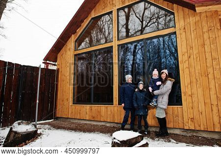 Attractive Mother With Her Kids In Winter Day Stand Near Wooden House With Large Window.